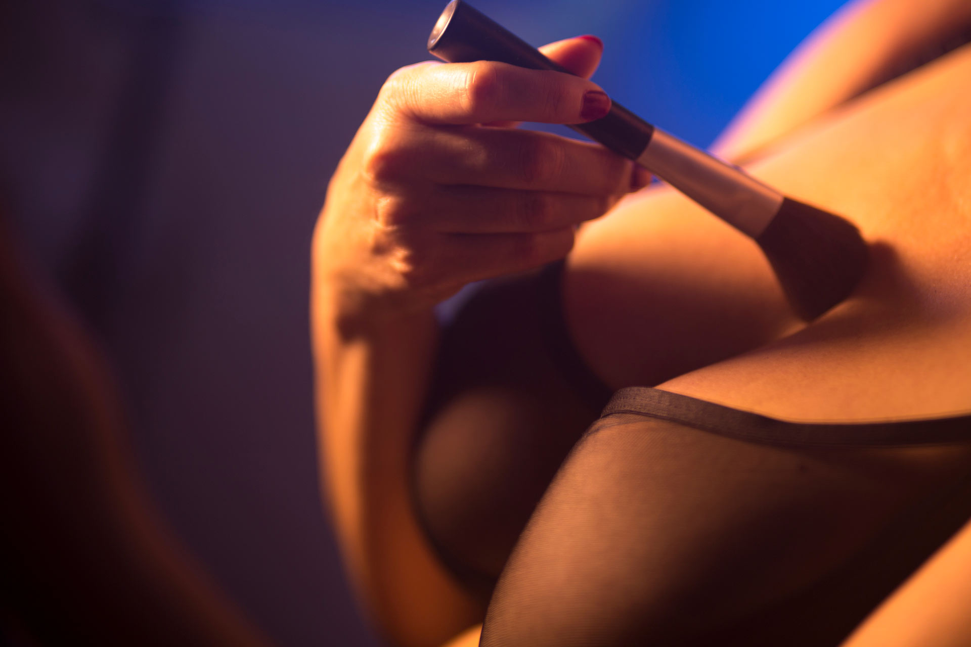 Cleavage of BBW woman in black bra holding a makeup brush and applying it on her chest, with blue light behind her.