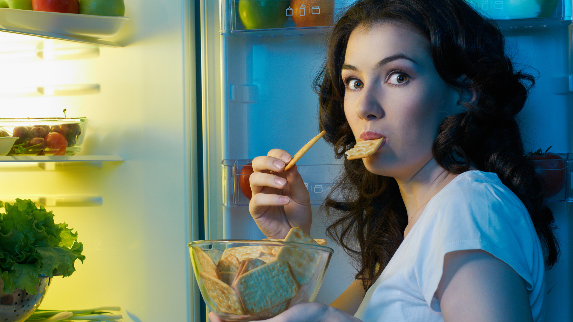 Brunette woman holding a bowl of graham crackers in front of an open fridge door, eating one while looking guilty.