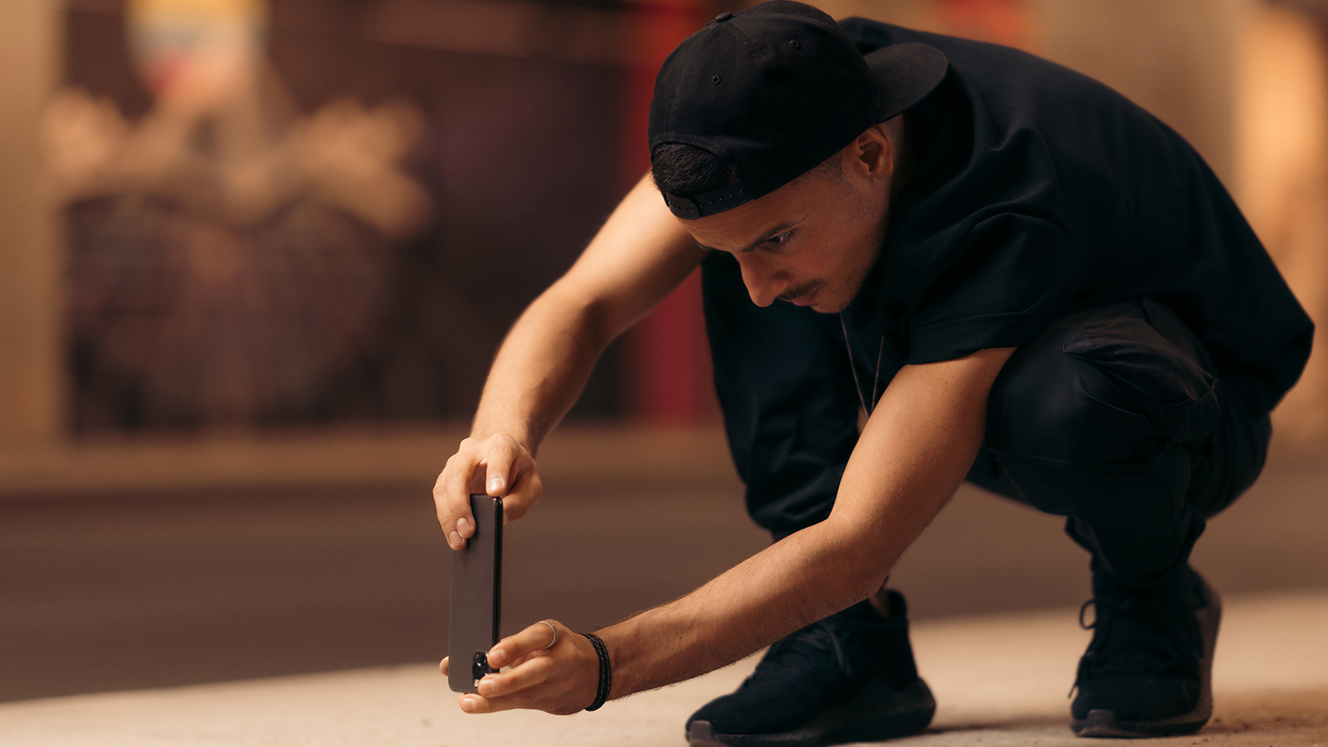 Man dressed in black shirt, trousers, shoes and hat, kneeling to hold his phone at a lower angle.