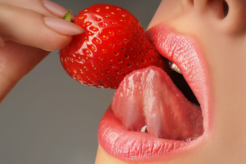 Female lips with open mouth showing teeth, and biting down on a strawberry tongue.