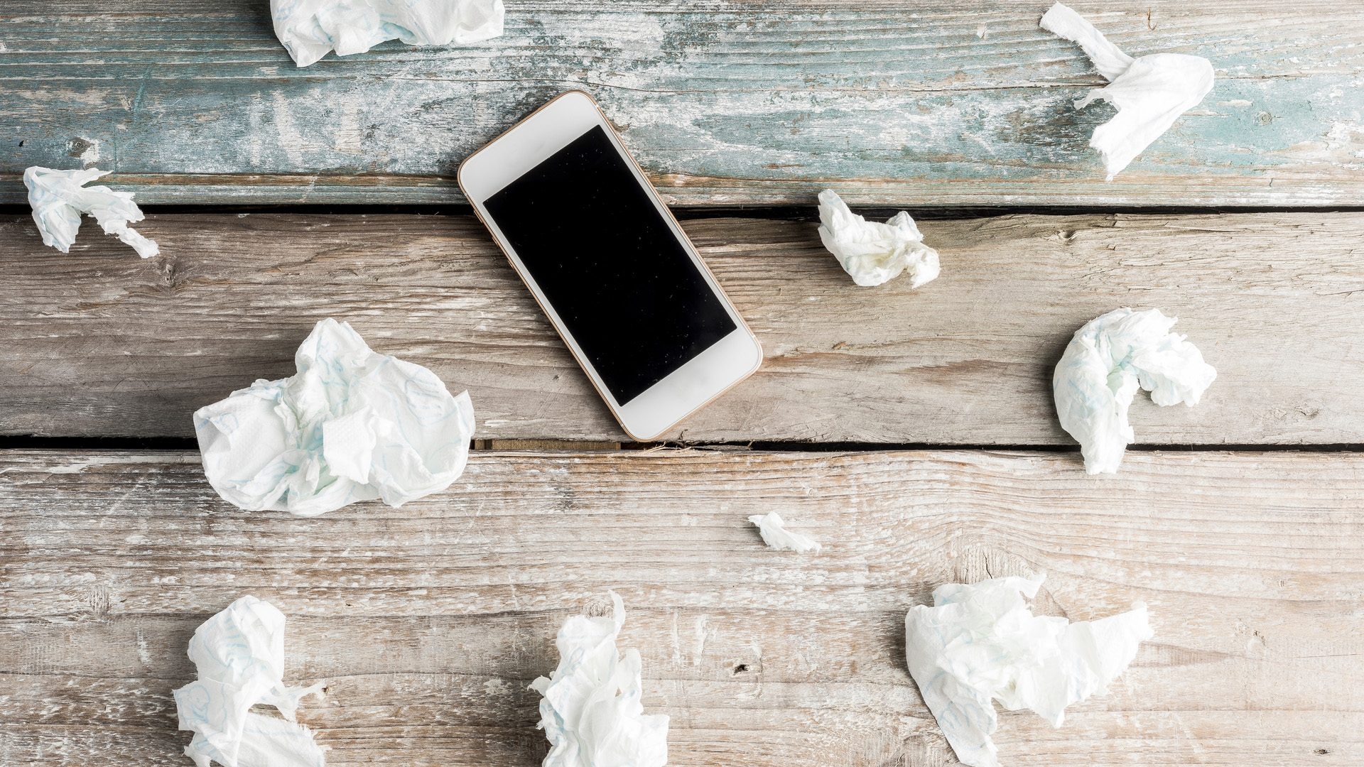 Mobile phone with black screen, on wooden surface surrounded by multiple crumpled up tissues.