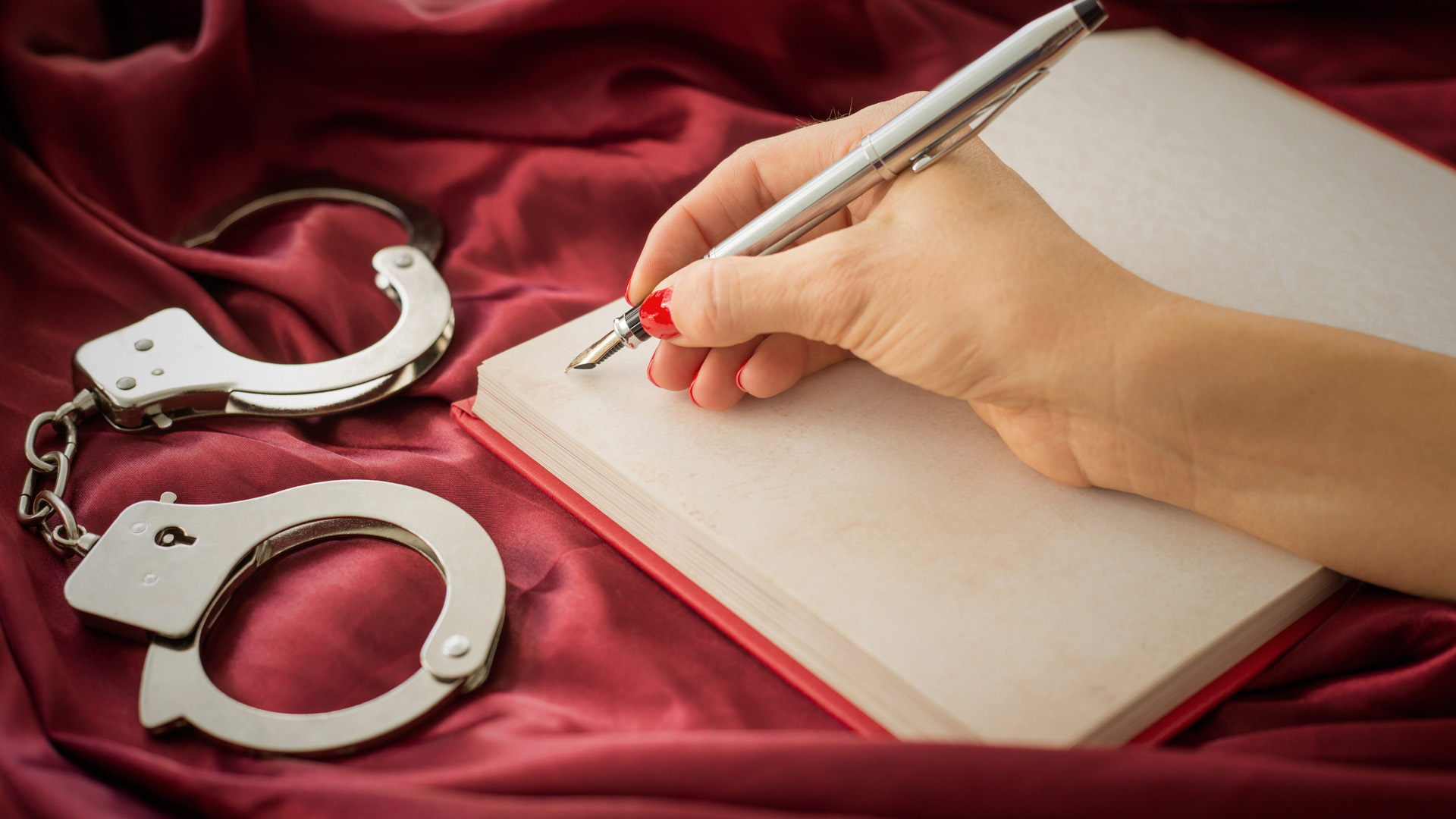 Hand of a woman holding a pen and writing on a text book with silver handcuffs next to it. Red silk background.